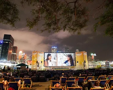 Outdoor cinema screening at night with city skyline and audience sitting on deck chairs.