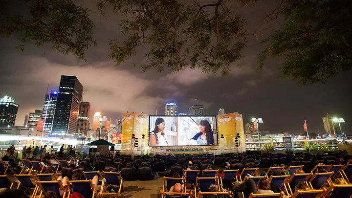 Outdoor cinema screening at night with city skyline and audience sitting on deck chairs.