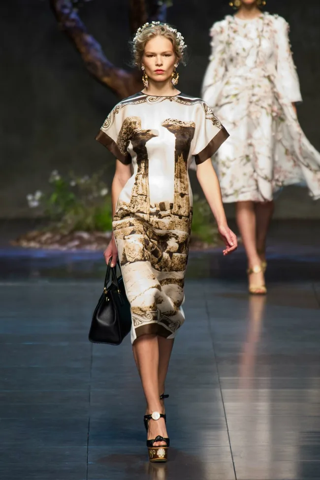 Model in Dolce & Gabbana SS14 runway wearing a dress with Greek ruins print, holding a black handbag.