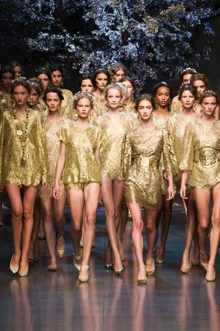 Models walking a runway in coordinated gold outfits, under a backdrop of flowers.