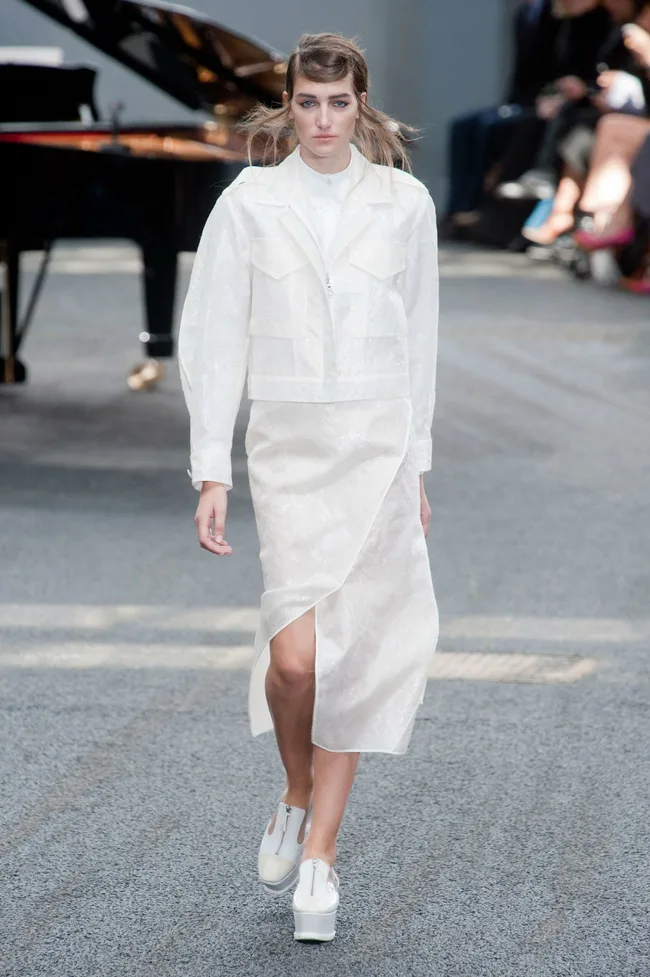 Model in a white outfit walking on a runway with a piano in the background at Erdem Spring/Summer 2014 fashion show.