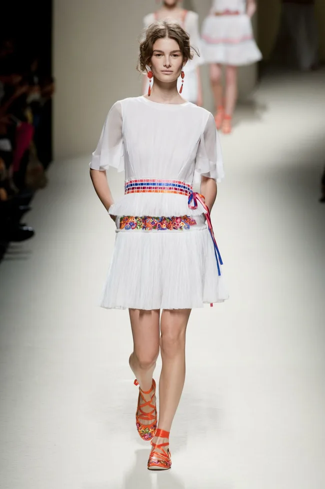 Model on runway in a white, flowy dress with colorful belt, orange sandals, and red earrings during Alberta Ferretti SS14 show.