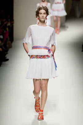 Model on runway in a white, flowy dress with colorful belt, orange sandals, and red earrings during Alberta Ferretti SS14 show.