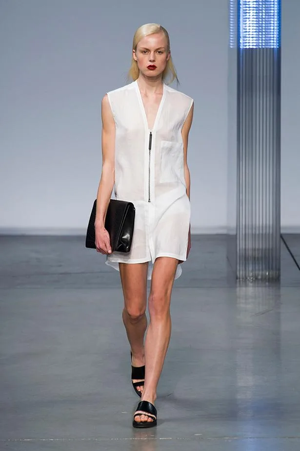 Model walking down the runway in a sleeveless white dress with a black clutch and sandals.