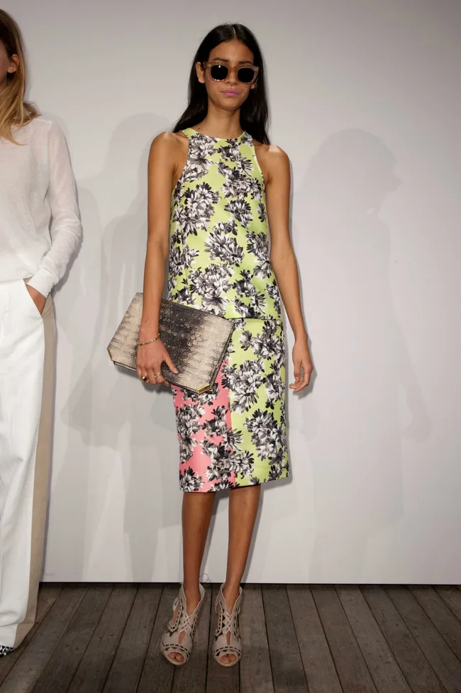 Woman in floral dress and sunglasses holding a clutch at a fashion show.