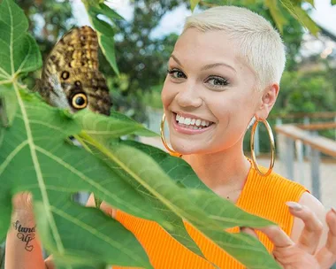 Jessie J smiles while holding large green leaves with a butterfly perched on them. She sports a short blonde haircut and hoop earrings.