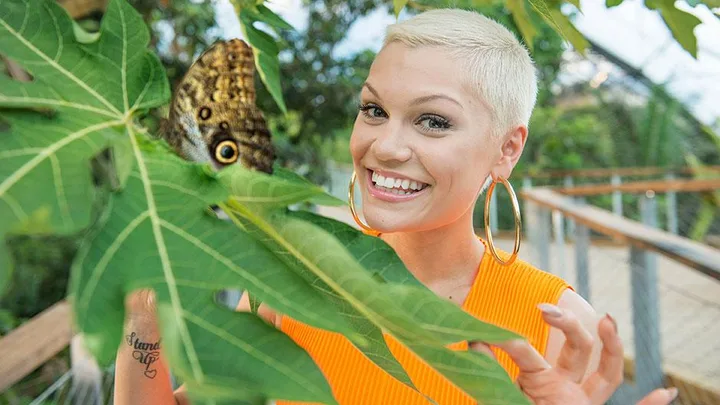 Jessie J smiles while holding large green leaves with a butterfly perched on them. She sports a short blonde haircut and hoop earrings.