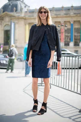 Woman in sunglasses wearing a navy dress and black jacket, standing outdoors with a blurred city background.