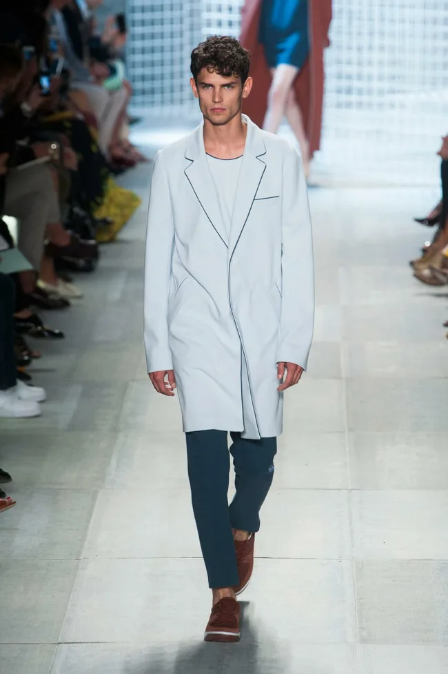 Model in a light blue coat and dark pants walking on a runway during Lacoste SS14 fashion show.