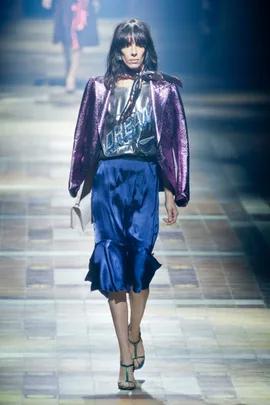 Model walks runway in Lanvin SS14: iridescent purple blazer, silver "Dream" top, blue satin skirt, holding white clutch.