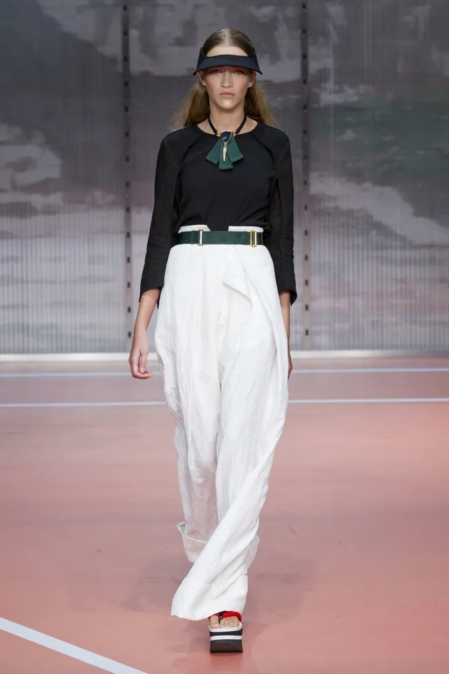 Model walking runway in black top, white high-waisted skirt, green belt, and visor, Marni SS14 collection.