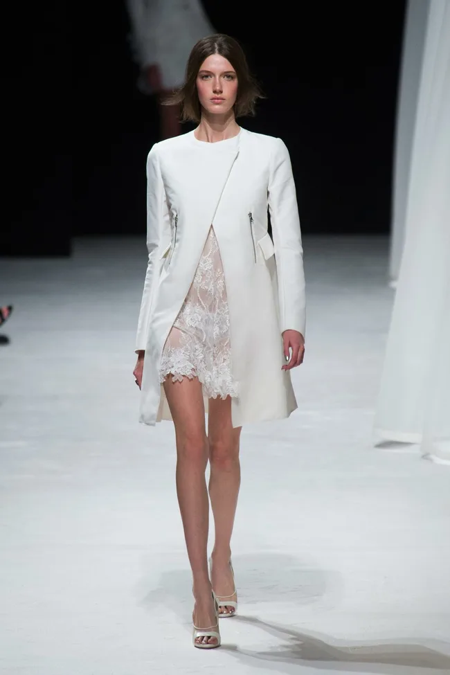 A model walks the runway in a white coat over a lace dress at the Nina Ricci Spring/Summer 2014 fashion show.