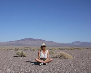 A person wearing a hat sits cross-legged in a dry, rocky landscape with mountains in the distance and clear blue sky above.