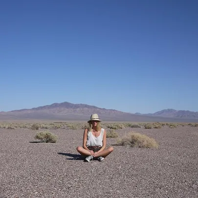 A person wearing a hat sits cross-legged in a dry, rocky landscape with mountains in the distance and clear blue sky above.