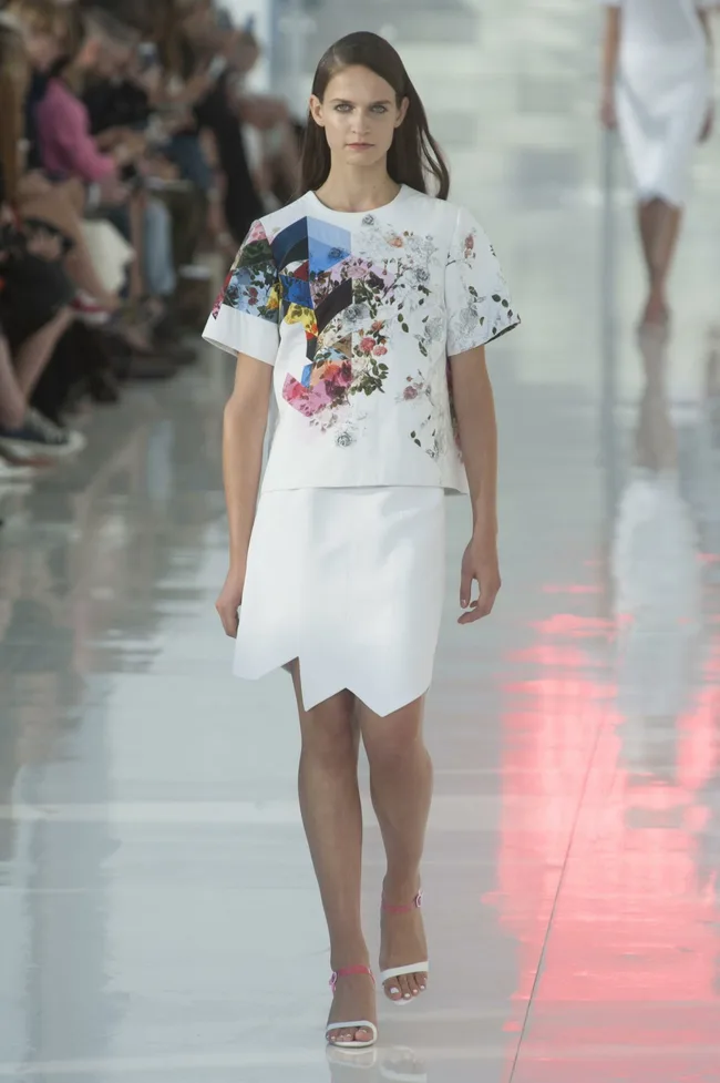 Model in a white floral top and white skirt walking on a runway with a seated audience in the background.