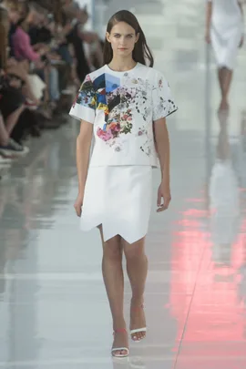 Model in a white floral top and white skirt walking on a runway with a seated audience in the background.