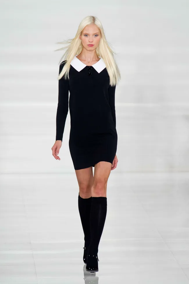 Model in a black dress with a white collar and knee-high socks walks on a bright runway.