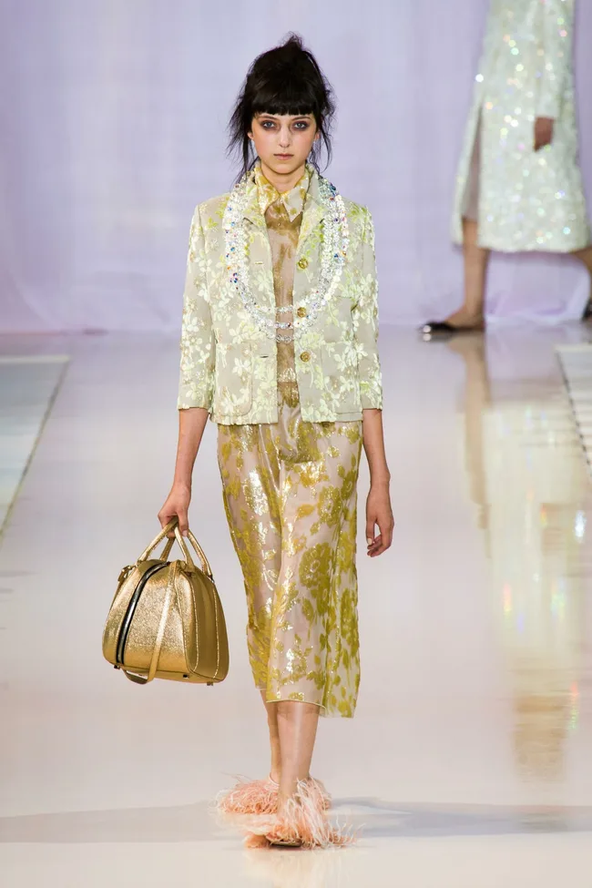 Model walks runway in sheer floral outfit with gold handbag and fluffy slippers at Rochas SS14 fashion show.