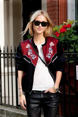 Woman in sunglasses wearing a floral bomber jacket, white top, and leather pants, standing outdoors.