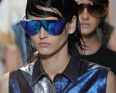 Model on runway wearing metallic blue top and large blue reflective sunglasses at fashion show.