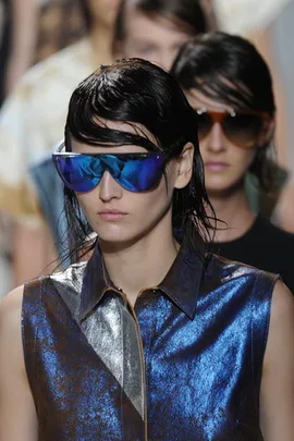 Model on runway wearing metallic blue top and large blue reflective sunglasses at fashion show.