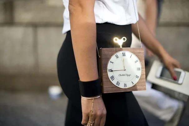 Woman with clock-shaped bag featuring Roman numerals, wearing a white top and black pants, holding a phone.