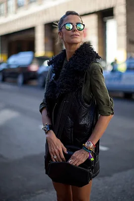 Woman at fashion week wearing reflective sunglasses, black fur vest, green top, and holding a bag.