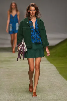 Model walking runway in green jacket, blue patterned blouse, green skirt, brown heels, holding a patterned bag.