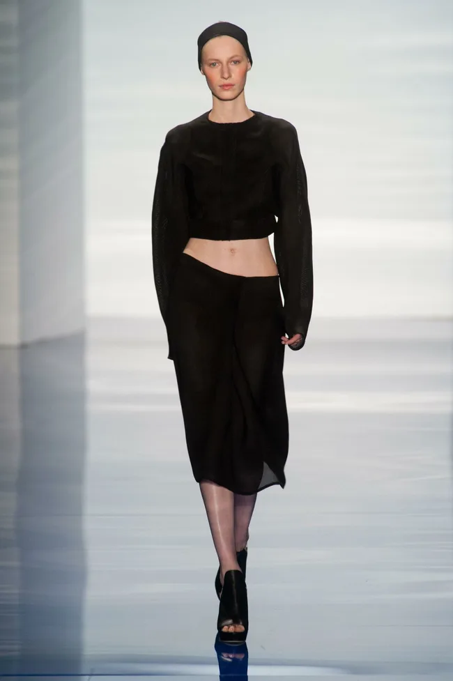 Model walking on a runway in a black, sheer outfit from Vera Wang's Spring/Summer 2014 collection.