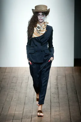 Model in a dark suit, straw hat, and scarf, with painted face, walking on wooden runway at Vivienne Westwood SS14 show.