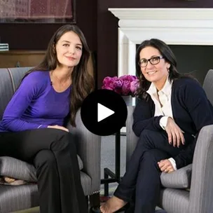 Katie Holmes and Bobbi Brown sitting together in armchairs, engaging in conversation, with flowers in the background.