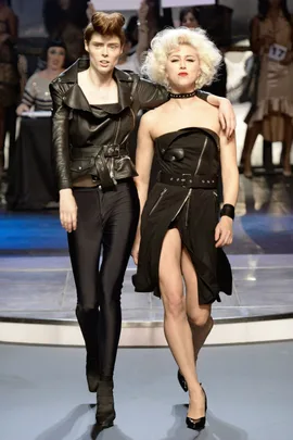 Two models on the runway; one in a black leather jacket and pants, the other in a strapless black dress with a high slit.