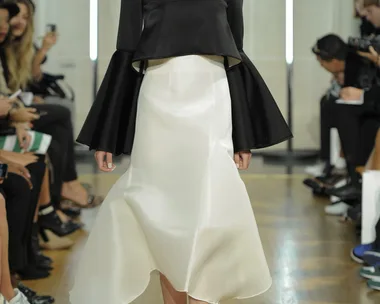 Model in a black top with flared sleeves, white skirt, and sparkly boots walks down a runway with audience on both sides.