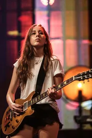 A member of the band Haim plays an electric guitar on stage in front of a colorful backdrop.