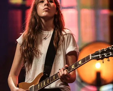 A member of the band Haim plays an electric guitar on stage in front of a colorful backdrop.