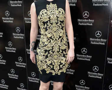 Woman in ornate black and gold dress at Mercedes-Benz Fashion Week backdrop.