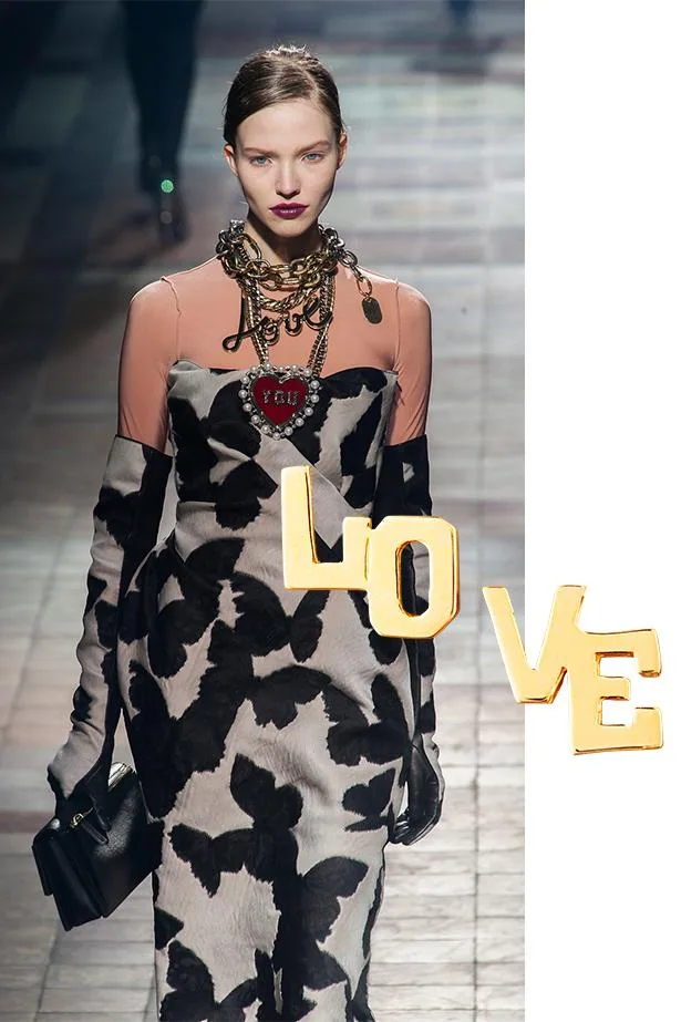 Model walking on runway in floral-patterned dress, wearing bold necklace, with "LOVE" in gold letters overlay.