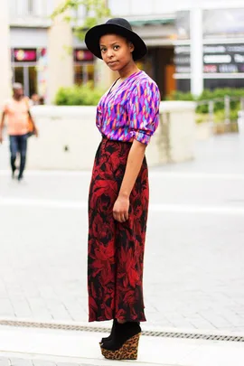 A woman in a vibrant blouse with patterns, red skirt, leopard print shoes, and a black hat on a street in South Africa.