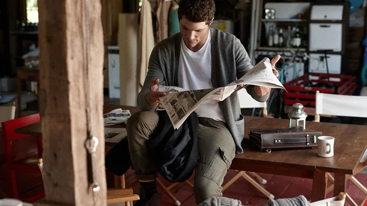 Man sitting in a cozy room, reading a newspaper while holding a jacket, with a briefcase and coffee mug on the wooden table.