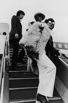 Three rock musicians in flamboyant outfits descend airplane stairs.