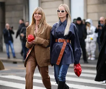 Two women walking in stylish jackets, one in brown with leather pants, the other in navy belted coat and sunglasses.