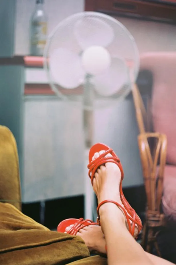 Feet in red sandals rest on a sofa; a white fan and a chair are in the background.