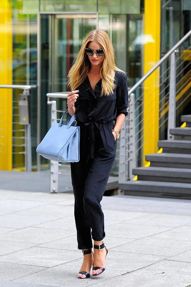 Person in black outfit and sunglasses walks with a blue bag near modern building with yellow accents.