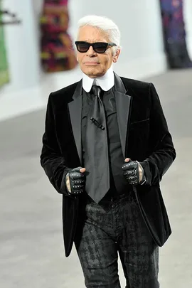 A man with white hair, black sunglasses, and black attire walks confidently indoors, hands in studded black gloves.