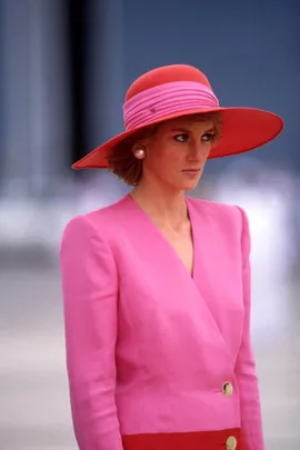 Princess in bold pink suit with matching wide-brimmed hat, looking serious at an outdoor event.