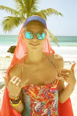 Person in colorful beach attire and sunglasses smiles in front of a palm tree and ocean backdrop.