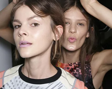 Two women posing playfully, one making a peace sign behind the other. Bright lighting and patterned clothing.