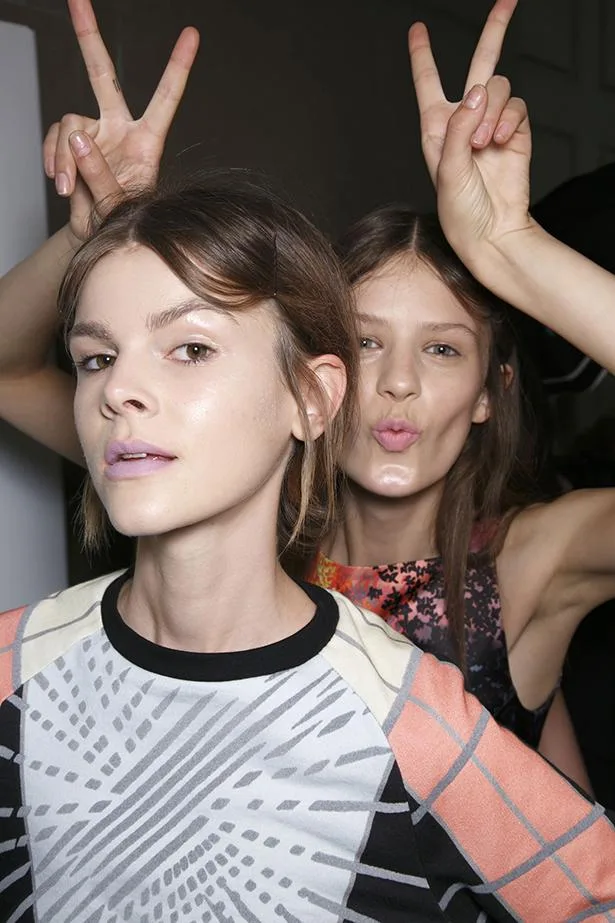 Two women posing playfully, one making a peace sign behind the other. Bright lighting and patterned clothing.