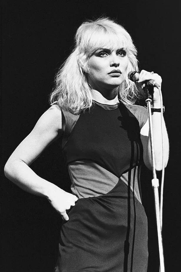 Blonde woman in a black dress holding a microphone on stage, looking determined.