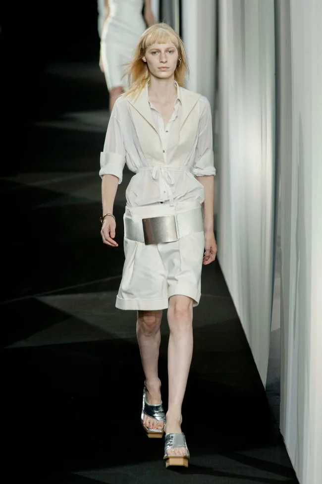 Model in white belted dress walks runway, paired with shiny sandals, during a fashion show.